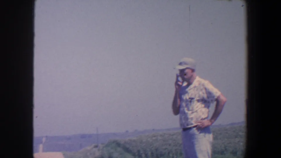 Adult man in rural Iowa stands by open farmland, one hand near his face and the other at his hip.