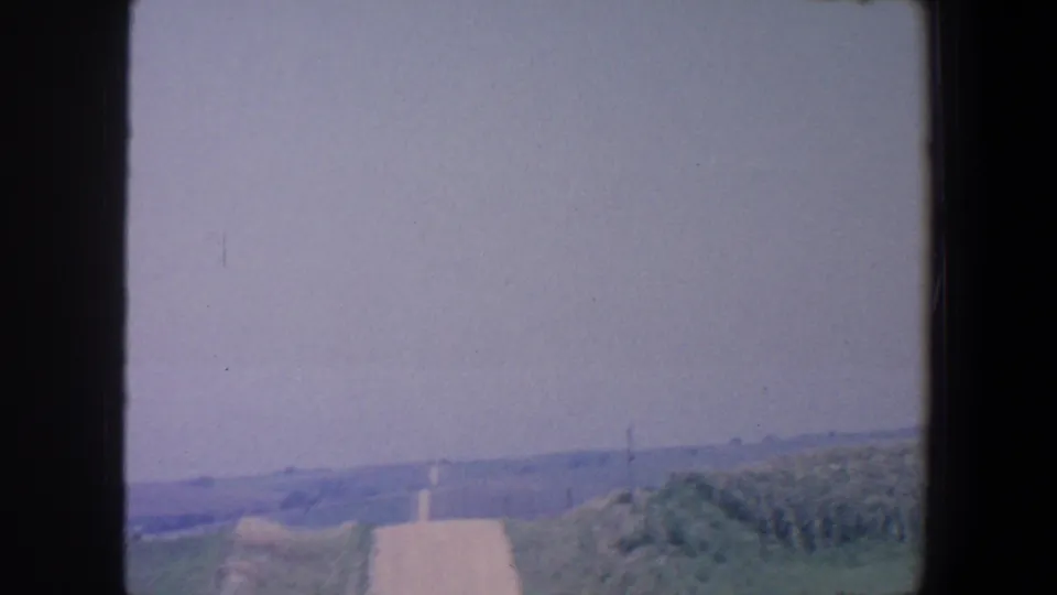 Rural Iowa landscape view shows open fields, a pale road or strip, distant posts, and a hazy sky.