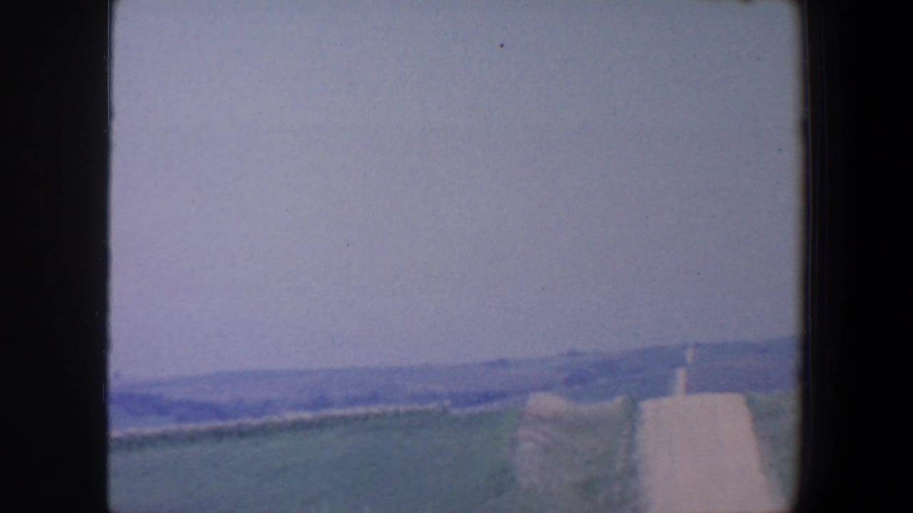 1959 Iowa countryside seen from a moving viewpoint, with open fields, a road edge, and hazy hills beyond.