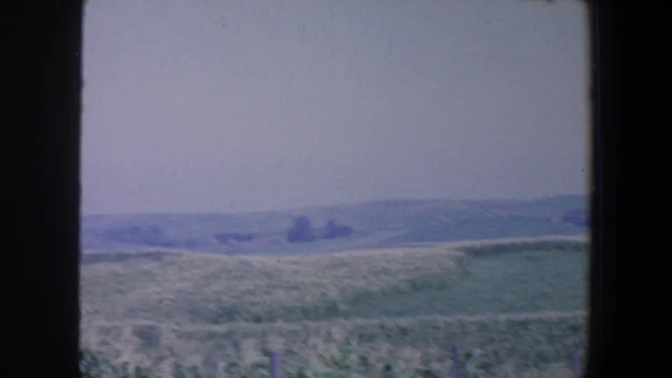 1959 Iowa rural landscape with green fields, low hills, and a hazy tree line in a faded 8mm frame.