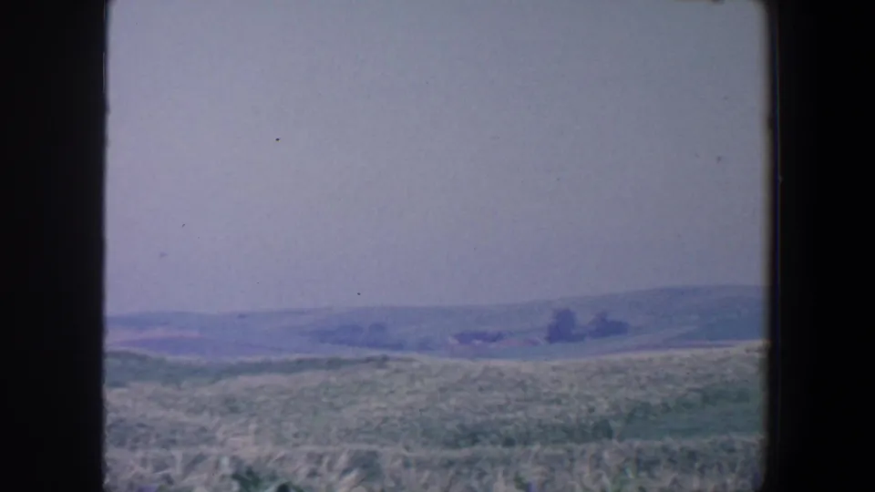 1959 Iowa countryside view with tall field growth, distant low hills, pale sky, and dark borders from the 8mm frame or window edge.