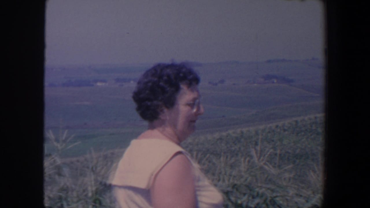 Woman in a light sleeveless dress stands beside a cornfield with rolling farmland behind her.