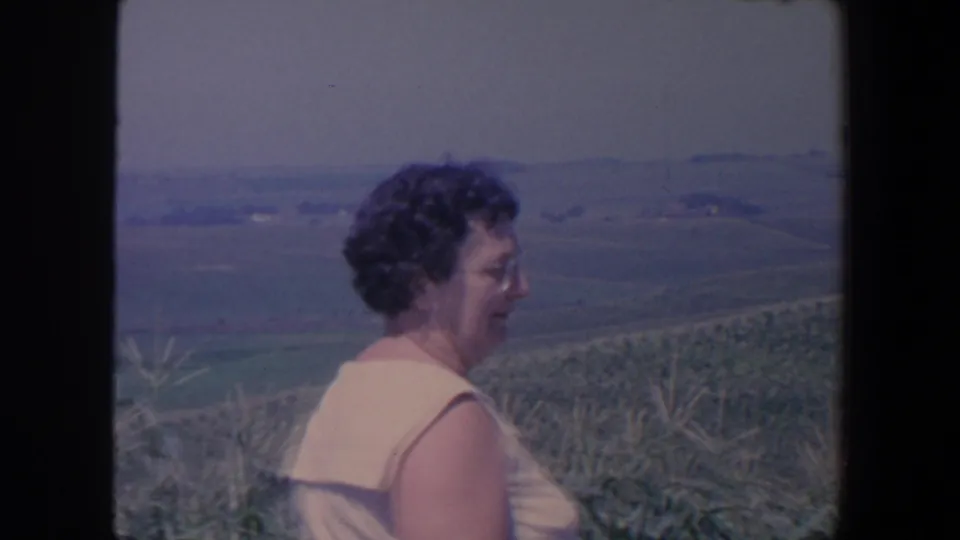 In Iowa home-movie footage from 1959, a woman in a light dress faces across a cornfield toward distant farmland and low hills.