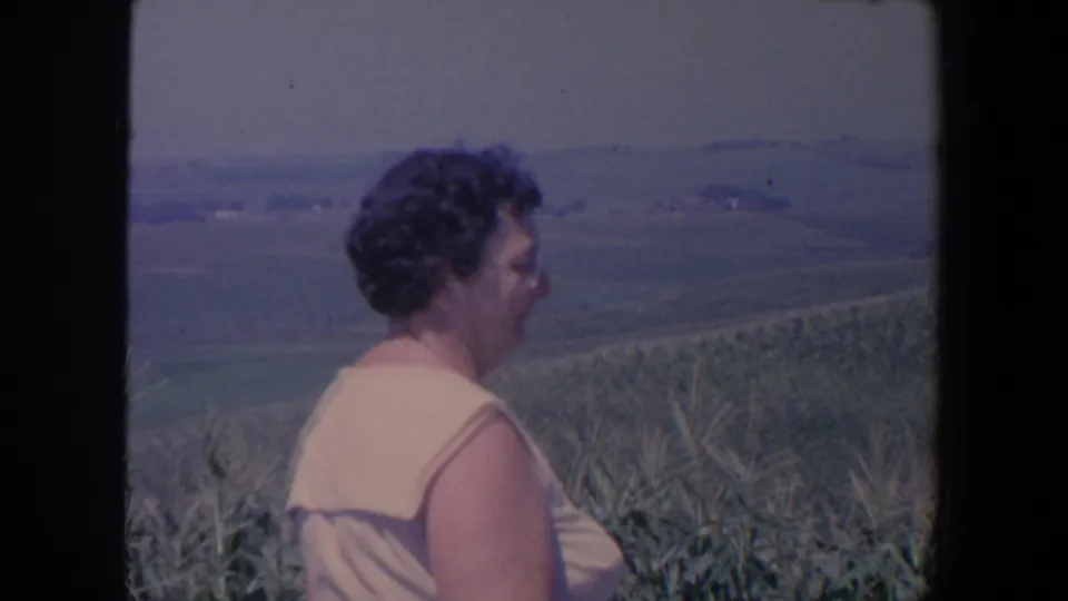 The woman remains in profile near tall corn, with soft 8mm color showing Iowa farmland and distant hills beyond the field.