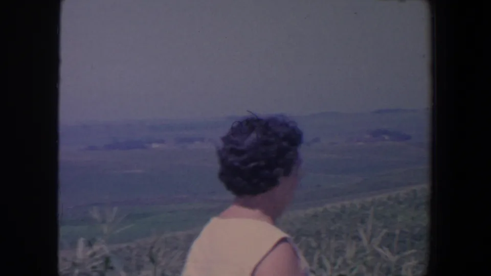 Seen from behind in the 1959 Iowa footage, the woman looks out over corn and rolling farmland under a pale sky.