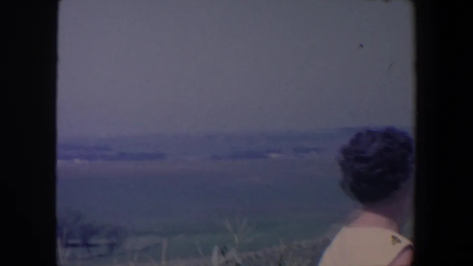 In the 1959 Iowa home-movie frame, a woman with curled hair stands on a grassy hillside facing a hazy rural landscape.