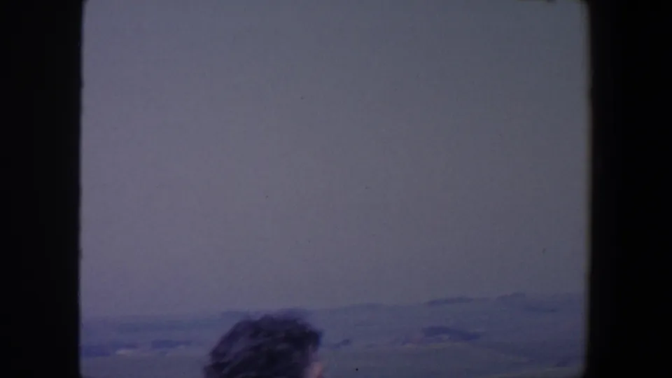The woman drops partly out of frame as the 1959 Iowa footage holds on hazy sky and distant rolling farmland.