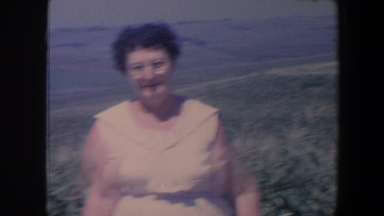 Woman in a pale pink sleeveless dress standing in a field with rolling farmland behind her.