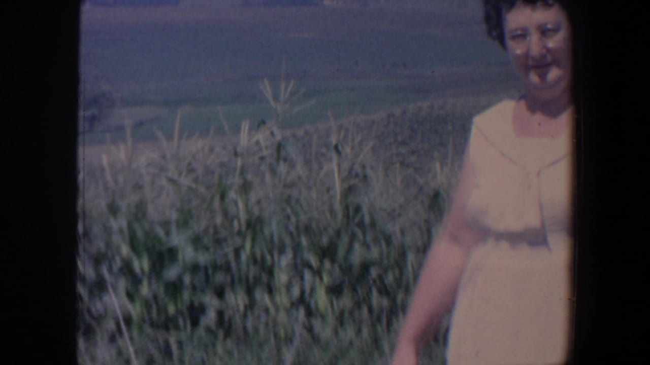 The woman stands near tall field plants in rural Iowa, with open farmland stretching into the background.
