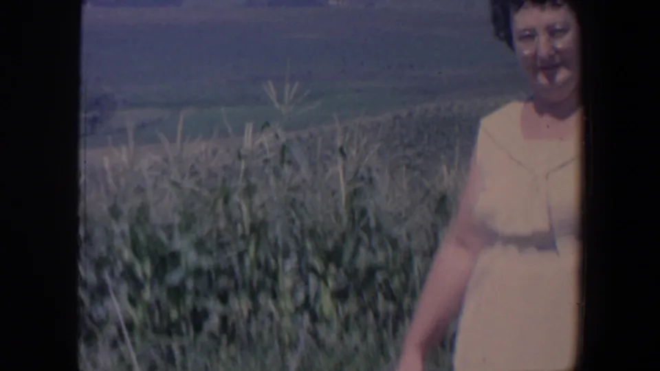 The woman stands near tall field plants in rural Iowa, with open farmland stretching into the background.