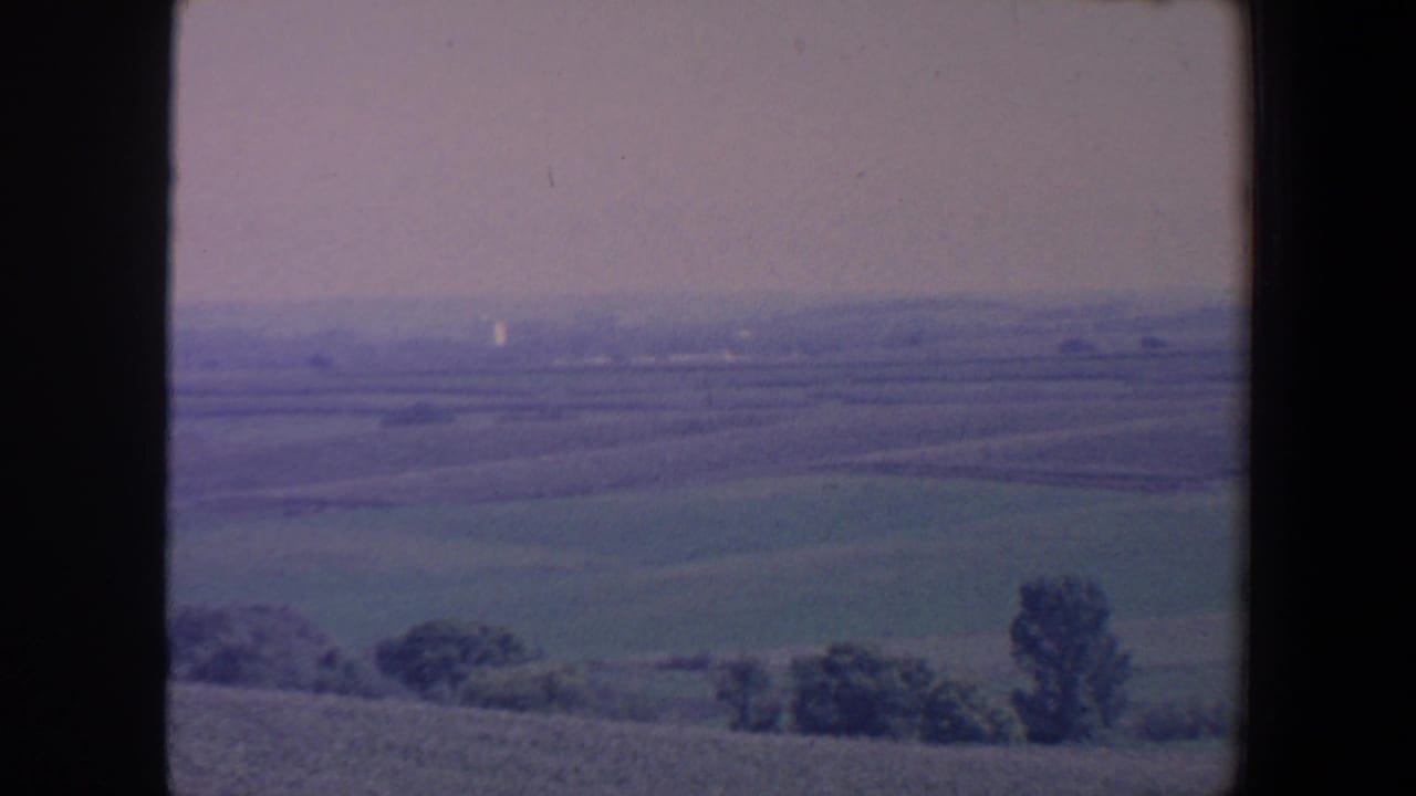 1959 Iowa home-movie frame with an elevated view across farmland, scattered trees and a hazy sky.