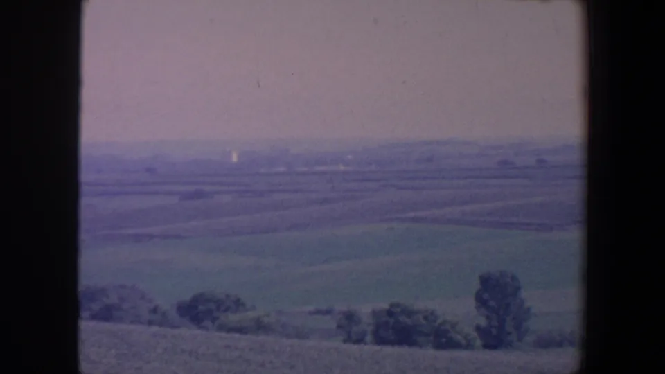 1959 Iowa home-movie frame with an elevated view across farmland, scattered trees and a hazy sky.