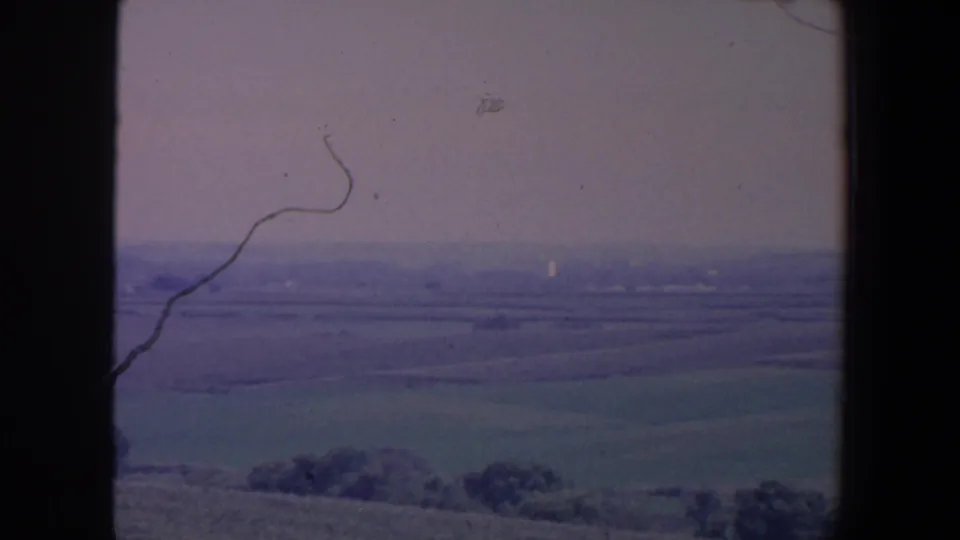 1959 Iowa frame showing broad fields, foreground trees and a hazy rural horizon in soft 8mm color.