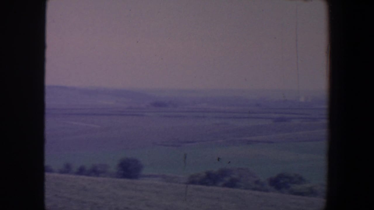 Rural fields and distant trees seen through a dark vehicle window frame.