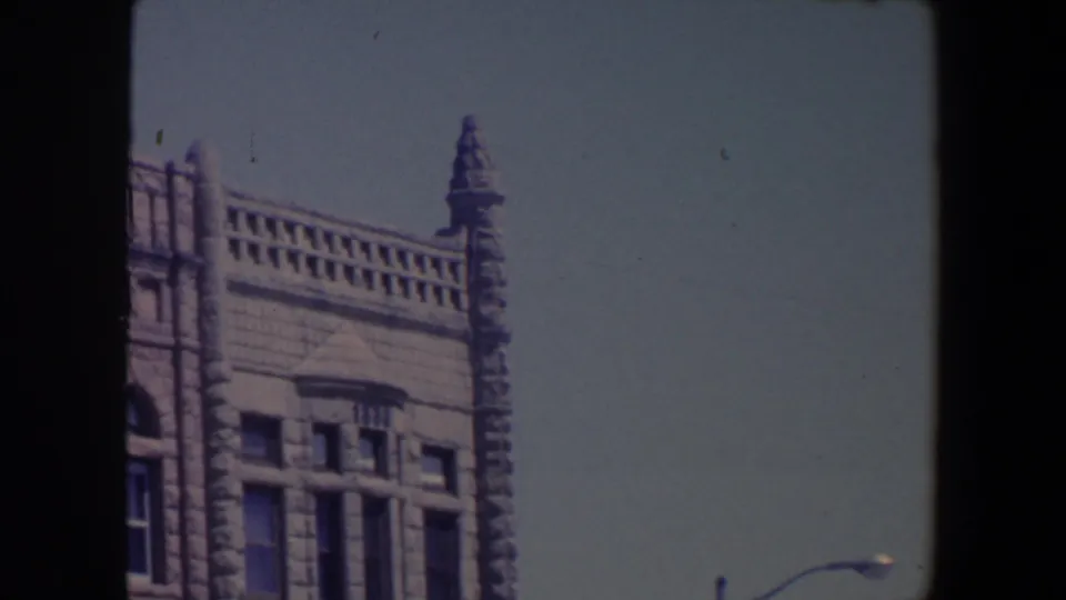 1959 Iowa 8mm view of a weathered stone facade with decorative trim, arched openings, upper windows, and a streetlamp below.