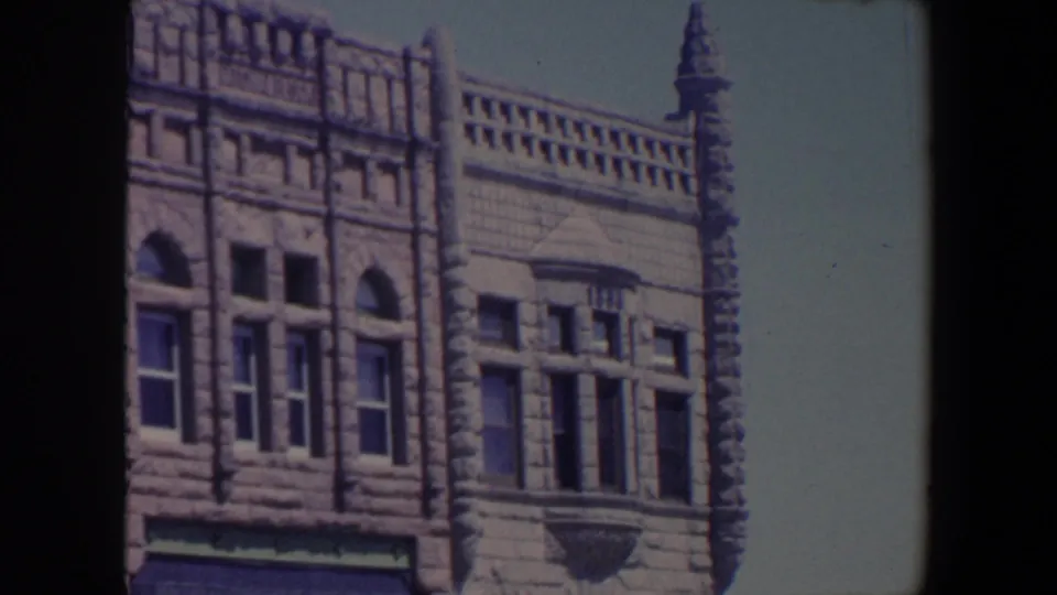 1959 Iowa frame showing a wider section of the ornate stone building, including arched windows, cornice work, spire, and pale sky.