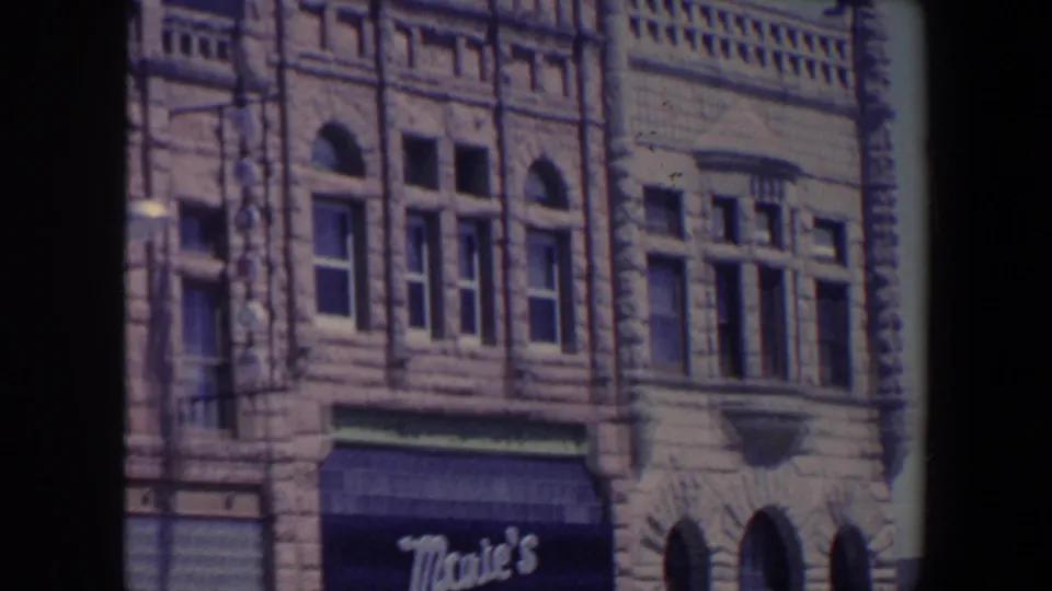 1959 Iowa home-movie frame showing an ornate stone storefront facade with arched windows and a dark awning reading Mae's.