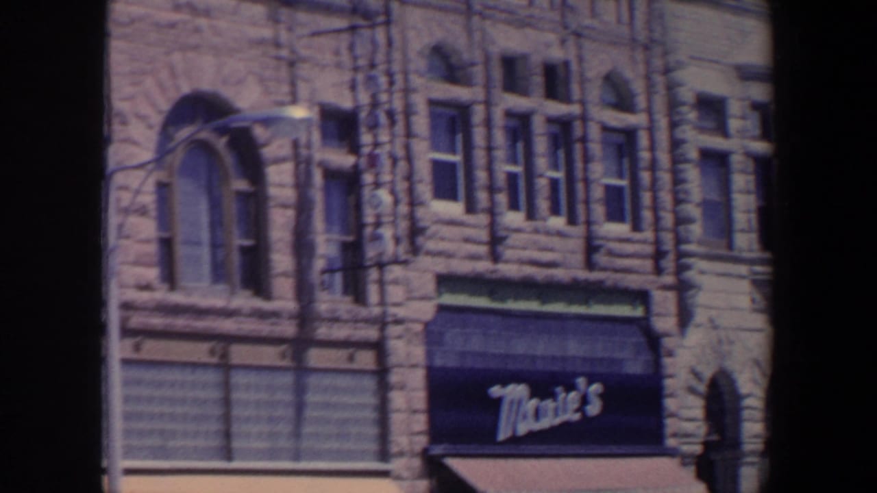 1959 Iowa frame with a stone commercial building, arched window, street lamp, and Mae's awning at street level.
