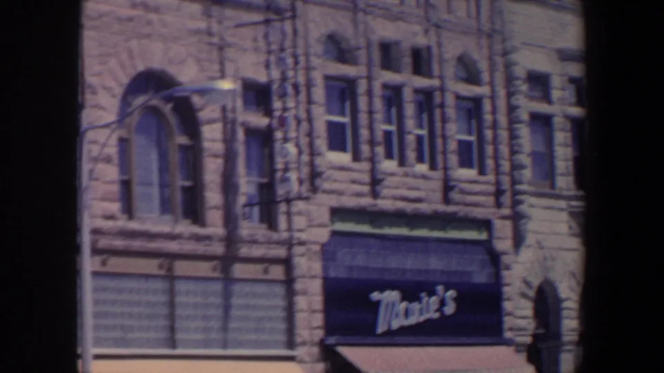 1959 Iowa frame with a stone commercial building, arched window, street lamp, and Mae's awning at street level.
