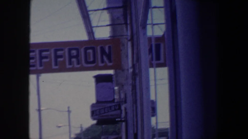 1959 Iowa home-movie frame showing part of a Heffron sign, storefront glass, and a smaller jewelry sign on a downtown street.