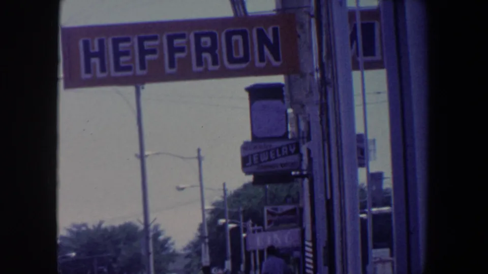 1959 Iowa street view showing a large Heffron sign, a jewelry shop sign, storefronts, trees, and streetlights in 8mm home-movie footage.