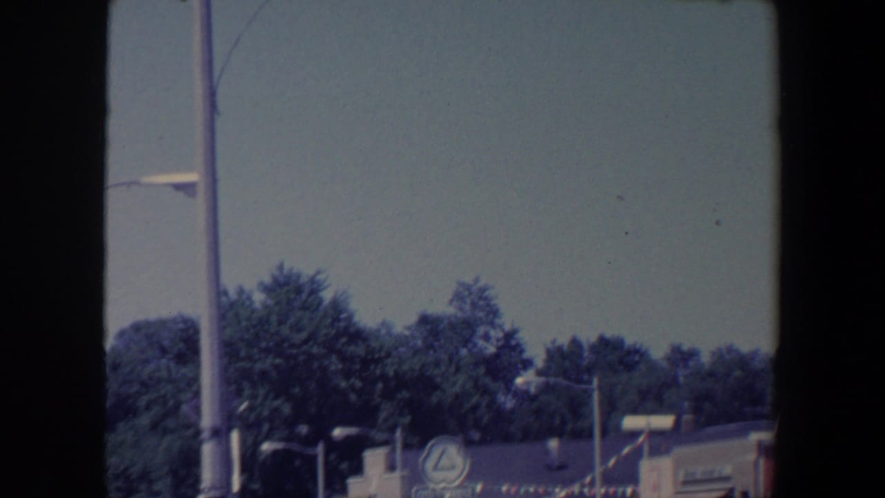 1959 Iowa street scene with treetops, streetlights, low storefronts, and a circular business sign near festive bunting.