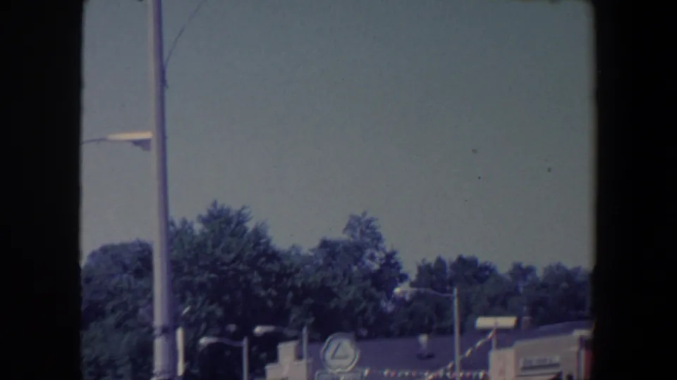 1959 Iowa street scene with treetops, streetlights, low storefronts, and a circular business sign near festive bunting.