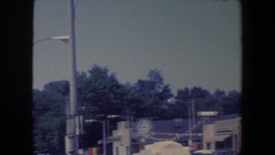 1959 Iowa storefront view showing festive bunting, streetlights, leafy trees, and a circular business sign in soft 8mm color.