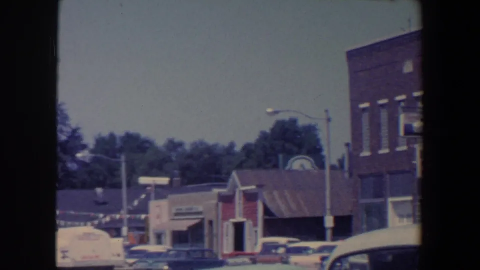 1959 Iowa downtown view with storefront awnings, parked cars, bunting, trees, and a brick building at the edge of frame.