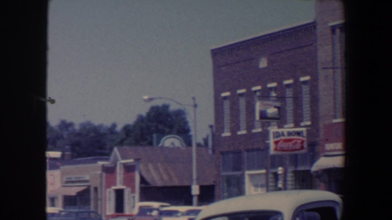 Small-town brick storefronts with parked cars, streetlights, and vintage signs.