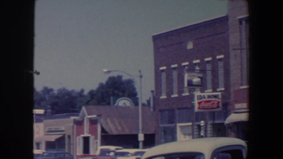 1959 Iowa street view with brick storefronts, parked cars, awnings, and visible Coca-Cola and shop signage.