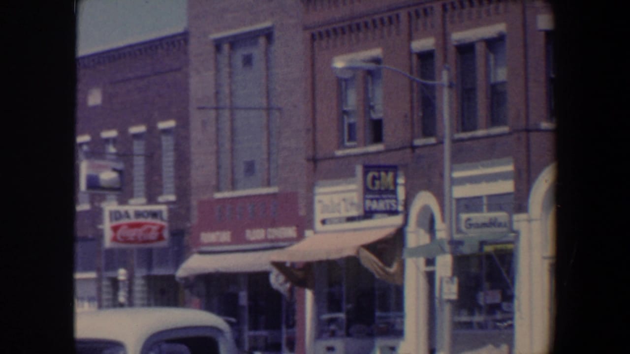1959 Iowa storefront row with awnings, a parked car in the foreground, and visible Coca-Cola and GM Parts signs.