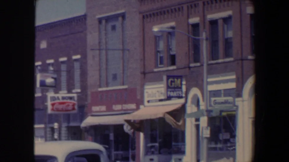 1959 Iowa storefront row with awnings, a parked car in the foreground, and visible Coca-Cola and GM Parts signs.