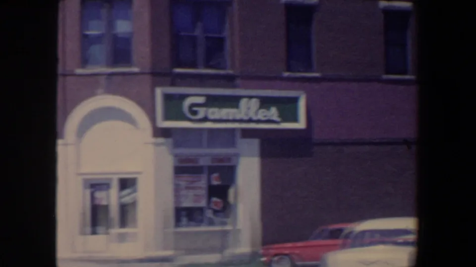 1959 Iowa street frame focused on a Gambles-style storefront, arched doorway, shop windows, and parked mid-century cars.