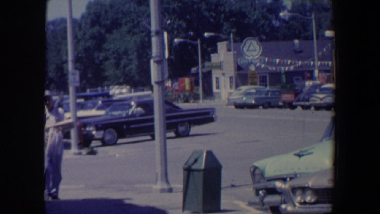 Small-town street with parked vintage cars, a walking man, utility pole, trash can, and storefronts.