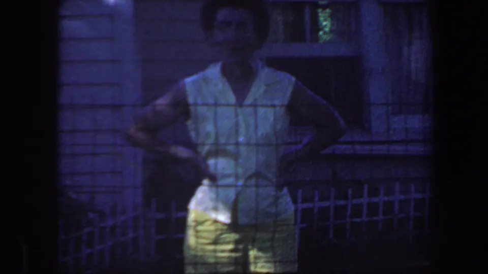 1959 Iowa home-movie frame showing a woman in a sleeveless top and yellow pants standing behind a wire fence near house siding and a window.