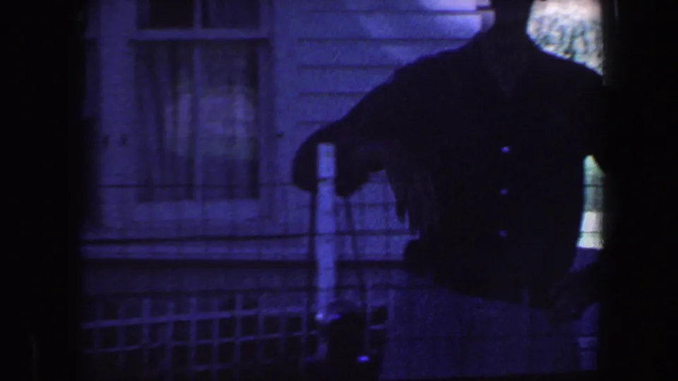 A man in a dark shirt stands in silhouette near the house and garden fence in blue-toned 1959 Iowa 8mm footage.