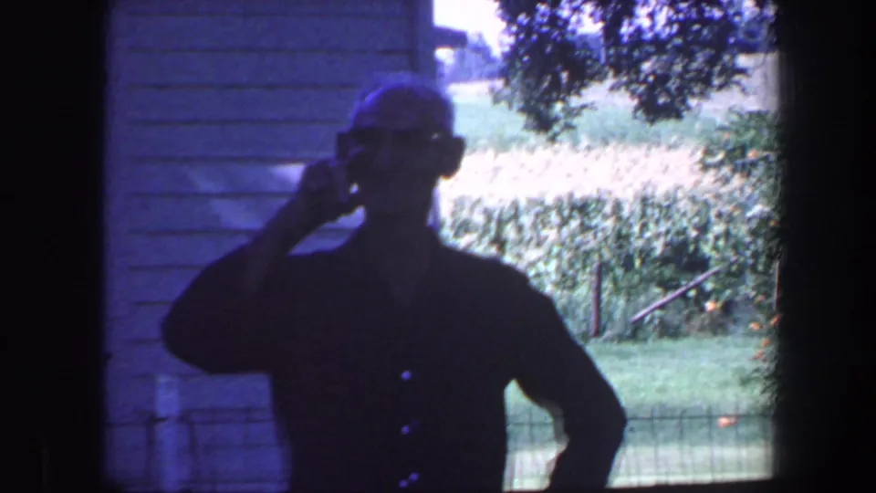 A silhouetted man raises his hand in a wave beside the house, with lawn, trees, and garden fencing visible in 1959 Iowa footage.