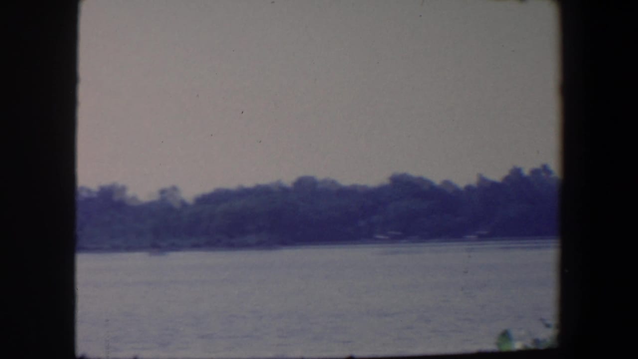 1959 Iowa home-movie frame with muted water, a distant wooded bank, pale sky, and dark film-gate edges.