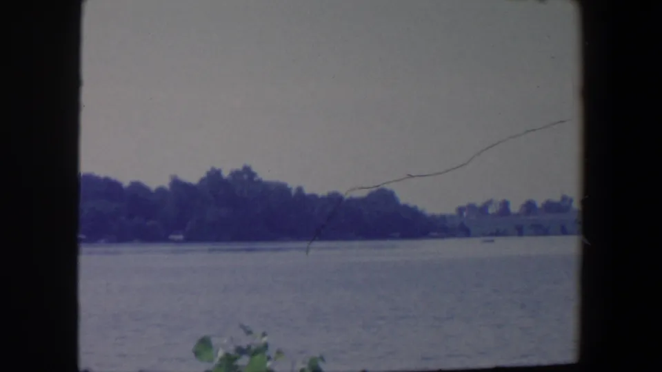 1959 Iowa 8mm frame showing open water, a tree-covered far shore, faint foreground leaves, and visible film wear.