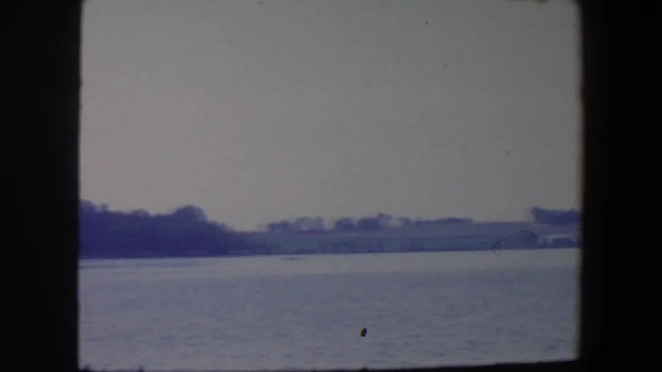 1959 Iowa home-movie frame with a wide expanse of water, low tree-lined horizon, pale sky, and soft blue-purple color shift.