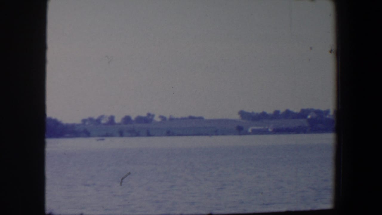 Calm water with a distant wooded shoreline and low hills under an overcast sky.