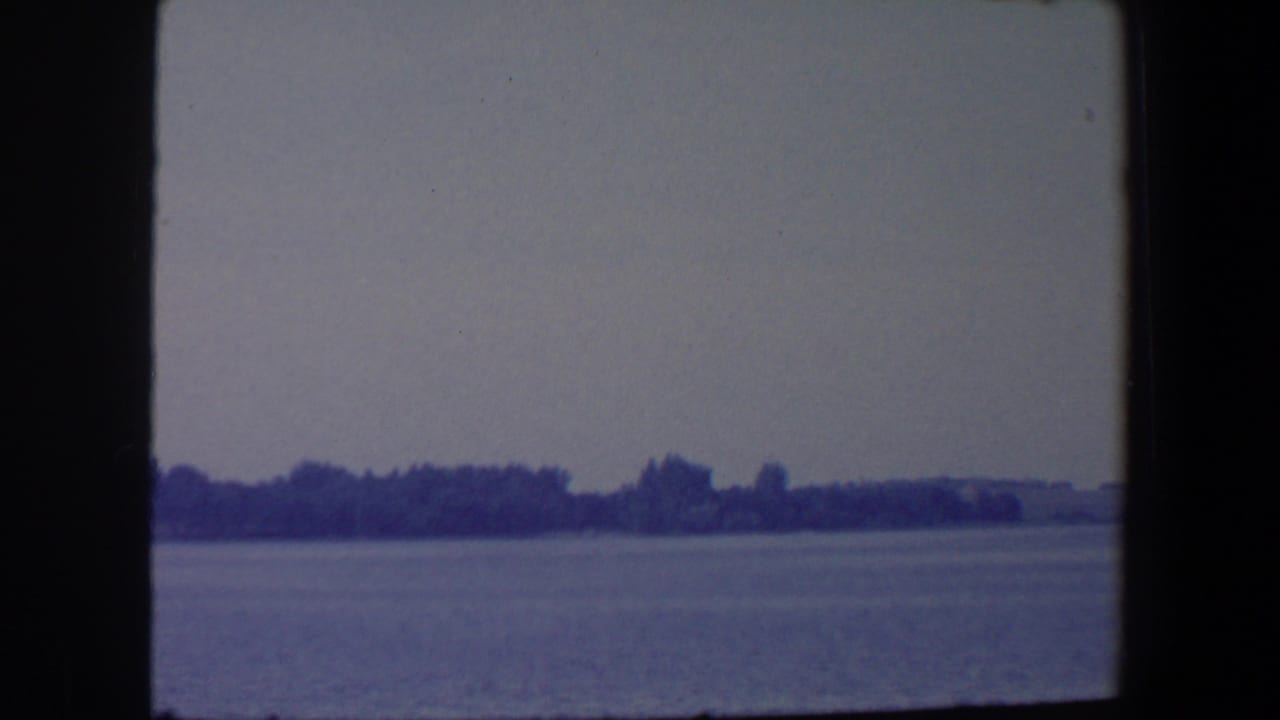 1959 Iowa home-movie frame with broad water in front of a dark tree line and low distant shore.