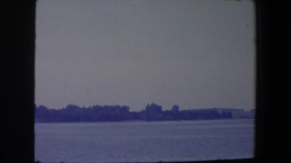 1959 Iowa home-movie frame with broad water in front of a dark tree line and low distant shore.