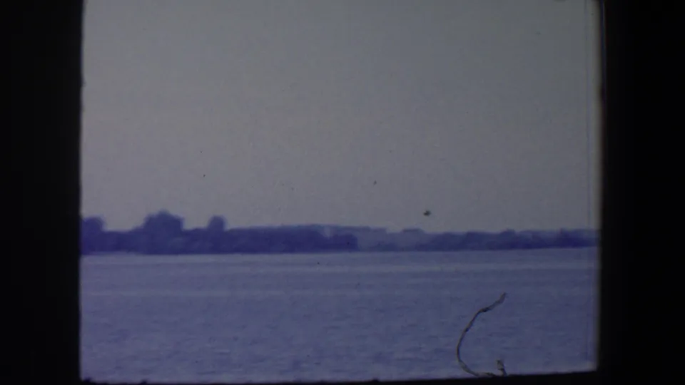 1959 Iowa 8mm frame showing water, a far rural shoreline, low hills, and a small bird or film mark above the horizon.