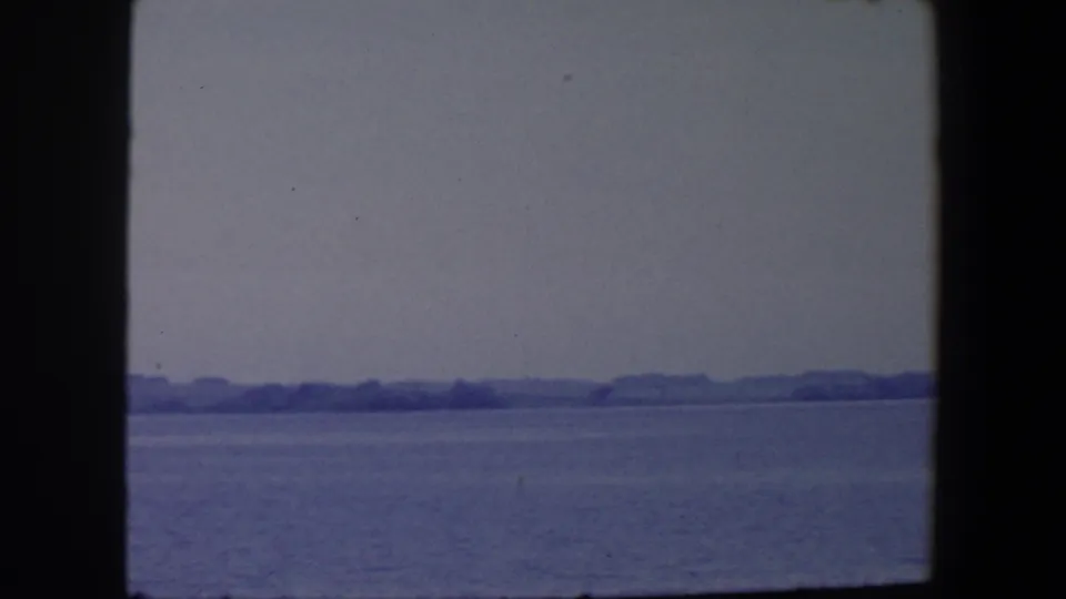 1959 Iowa home-movie frame of open water facing a faint, low wooded shoreline under an overcast sky.