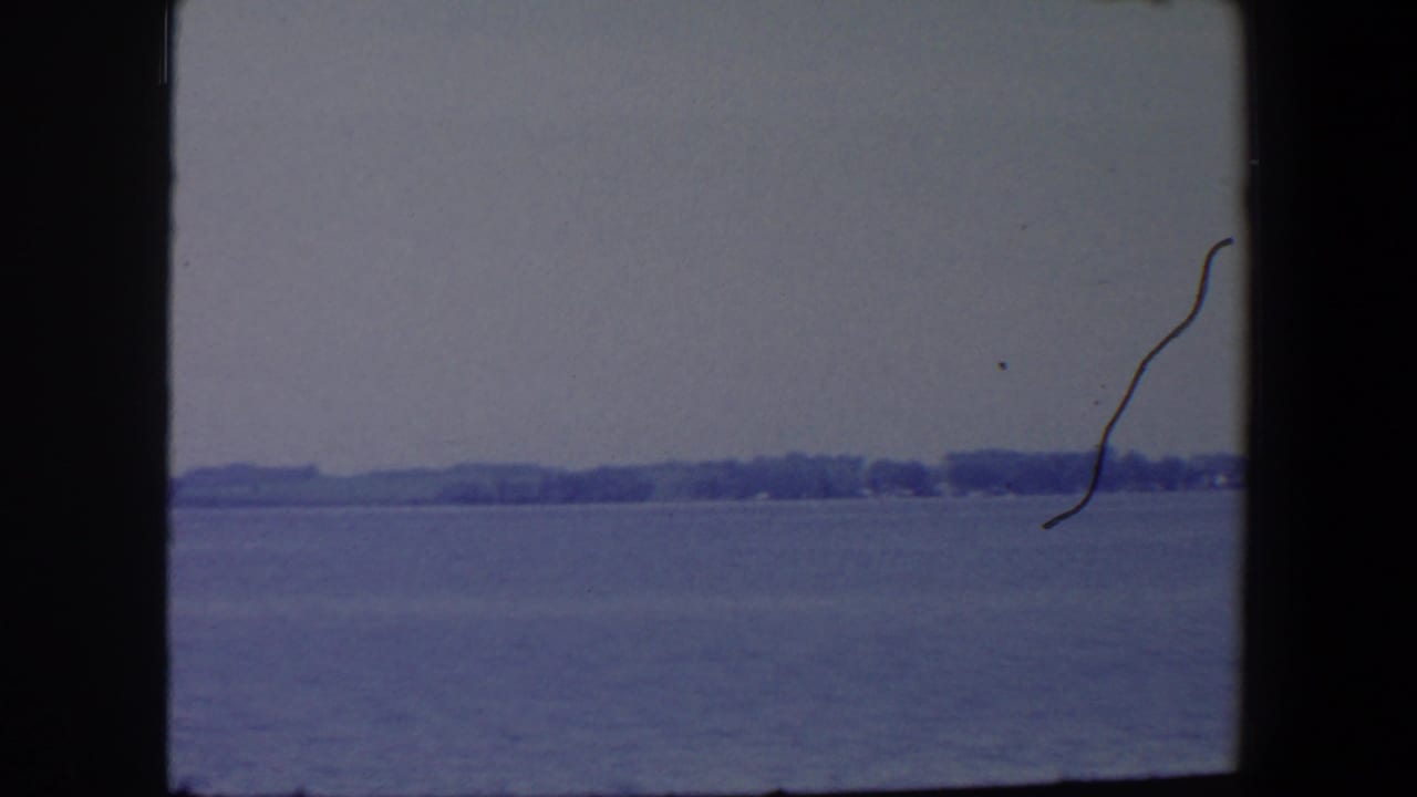 Wide view over still water toward a low wooded shore, with a dark curved film mark at right.