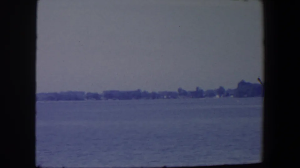 1959 Iowa 8mm frame showing calm water, a far tree line, and faint shoreline structures in soft focus.