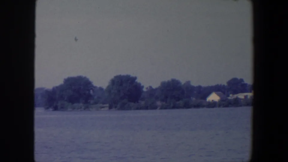 1959 Iowa lakefront view with still water, mature trees, and a small light-colored house near the shore.
