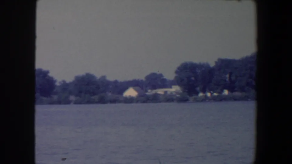 Soft 8mm frame of an Iowa lakefront with calm water, trees, and small houses set back from the shore.